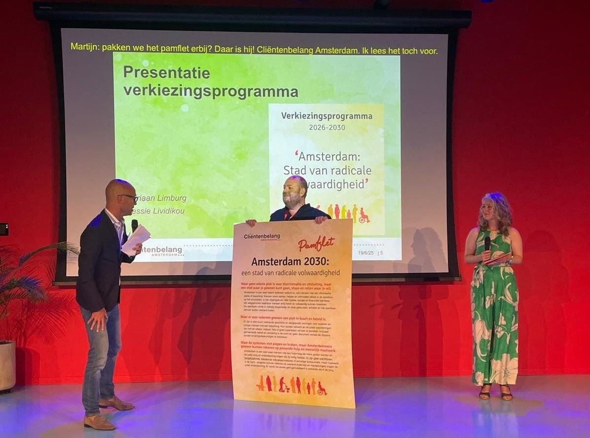 In een zaal staan dagvoorzitter Martijn de Greve, Jurriaan Limburg (beleidsmedewerker Ouderen) en Dessie Lividikou (beleidsmedewerker Wmo). Jurriaan houdt een groot bord vats met daarop de tekst van ons pamflet.